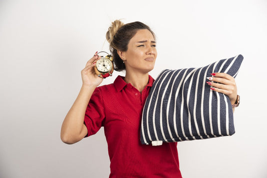 Woman in a red shirt holding a pillow and an alarm clock, looking annoyed.